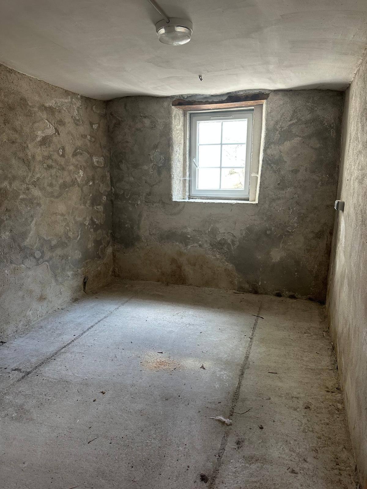 Pièce brute avant rénovation dans le Briançonnais : murs en pierre et béton décroûtés, sol en dalle nue et petite fenêtre à petits carreaux