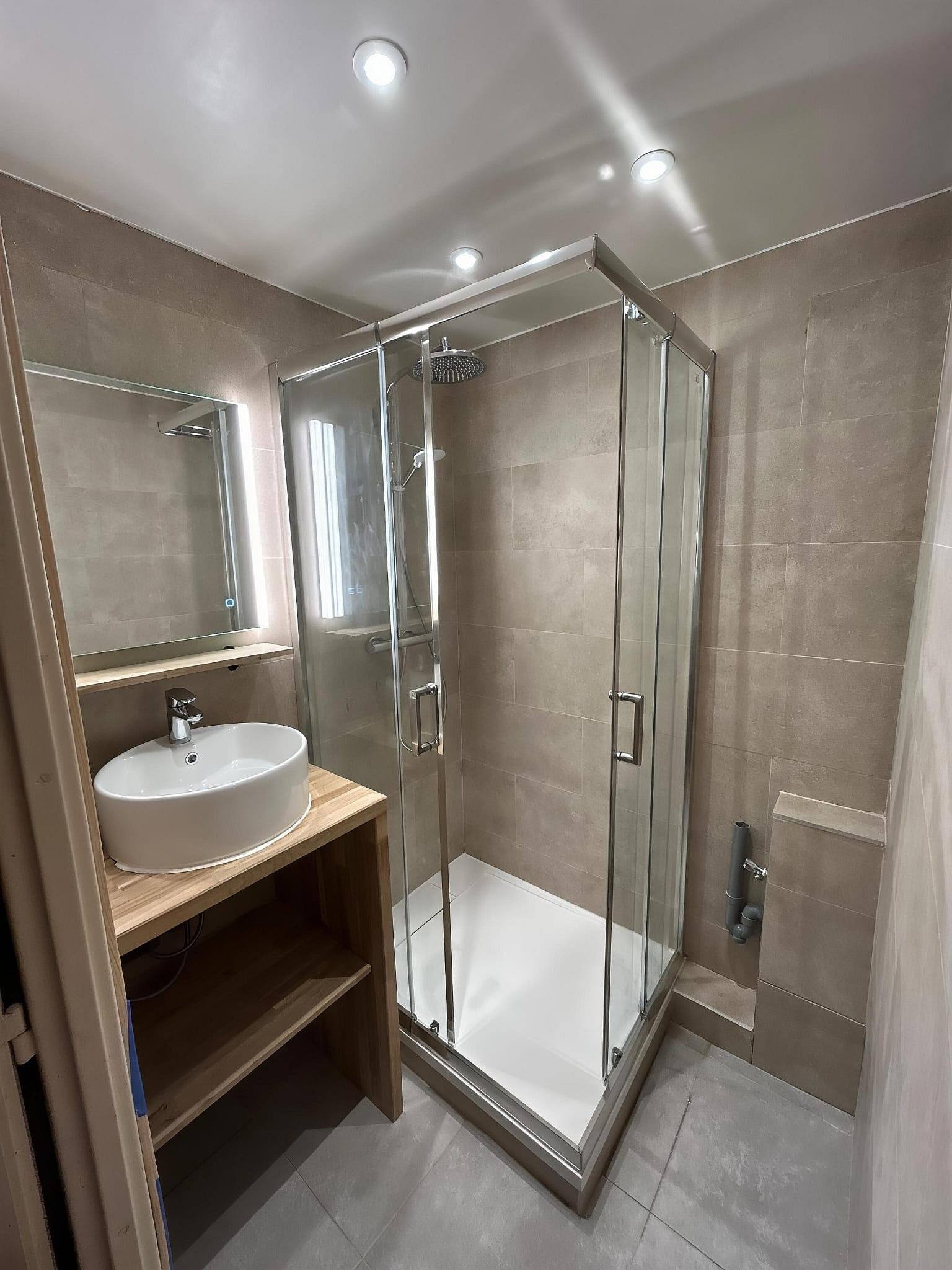 La même salle de bain après rénovation : douche à l’italienne, carrelage grand format effet pierre beige, vasque sur plan bois et miroir rétroéclairé