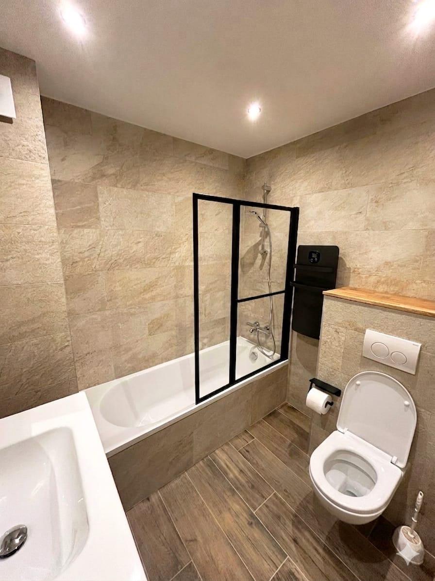 Salle de bain rénovée avec baignoire, paroi vitrée noire, carrelage effet pierre beige et WC suspendu