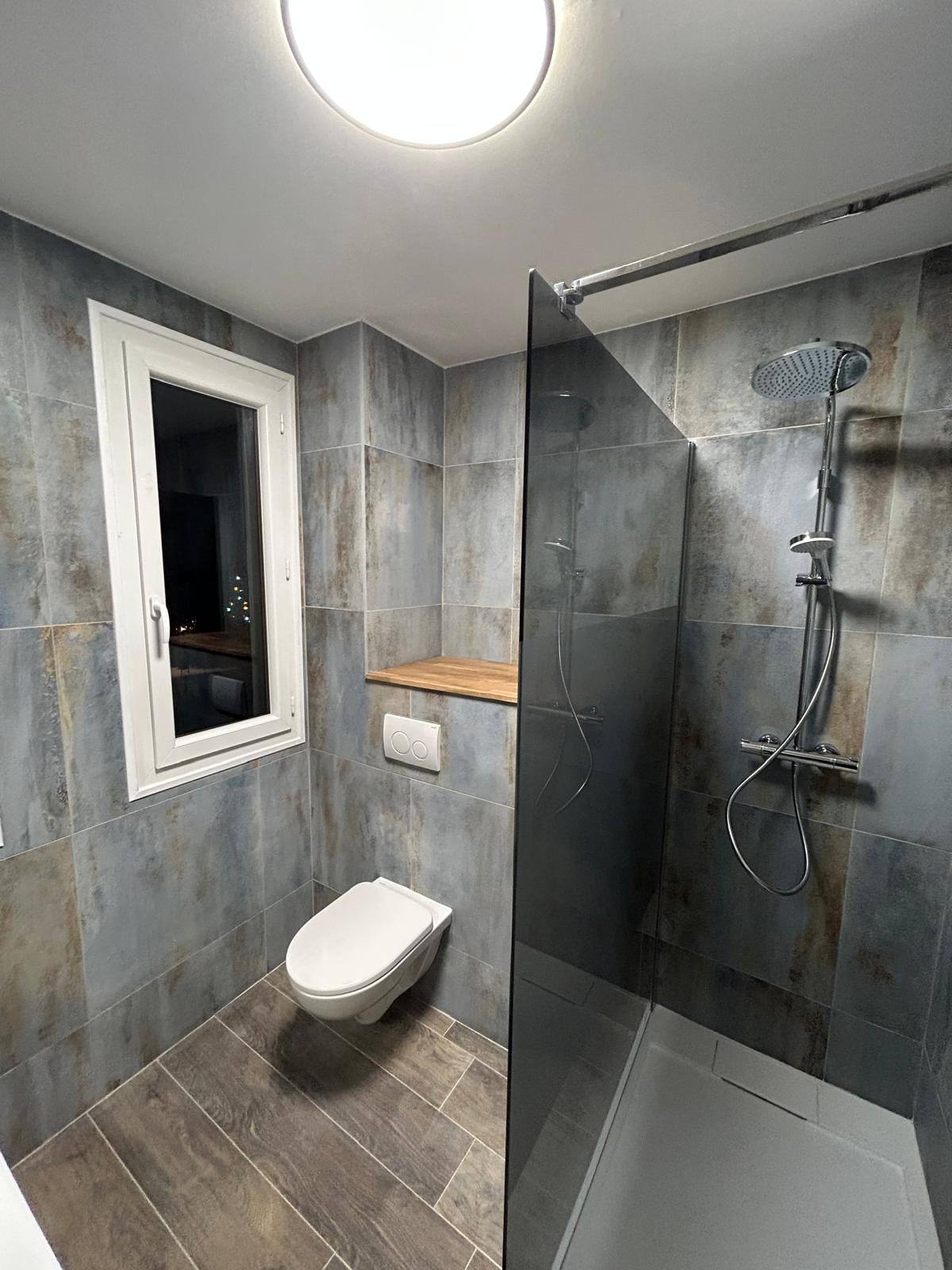 Salle de bain rénovée avec douche à l’italienne, carrelage gris effet métal, plan bois et WC suspendu