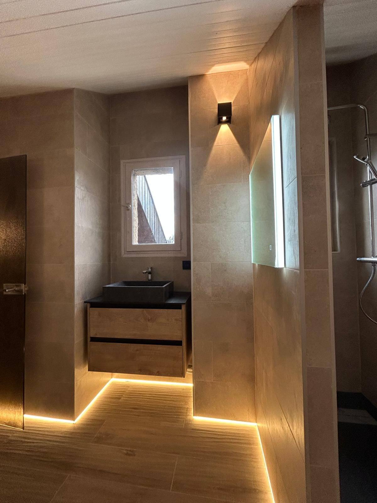 Salle de bain aménagée sous pente avec murs en béton ciré, vasque noire sur meuble suspendu, bandeau LED au sol et plafond en lambris blanc